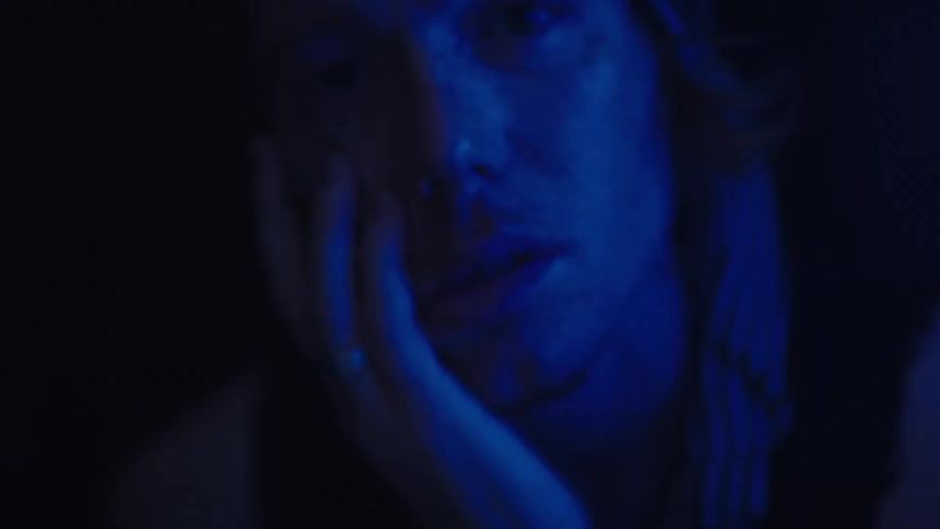 A closeup shot of a mans face in a dark room with blue lighting showing him with his hand touching his cheek and mouth as if in discomfort or pain