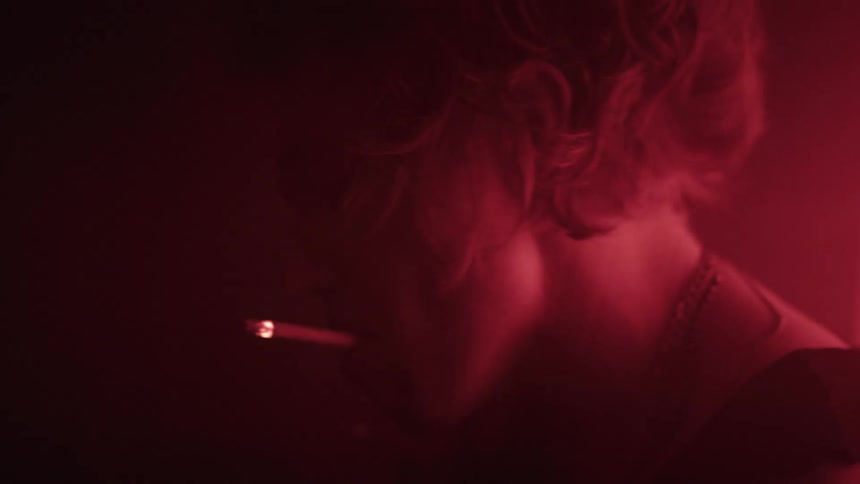 A man with long light colored hair and a chain necklace smokes a lit cigarette with his head tilted down and his face partially illuminated by a deep red light