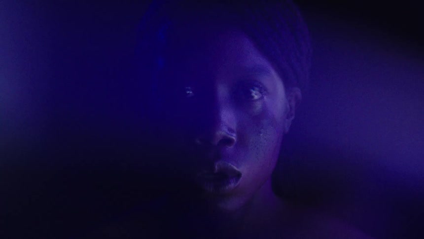 A young black woman sits in a dark room with light reflecting on her looking angry and sad then suddenly lowers her face as tears roll down her cheek