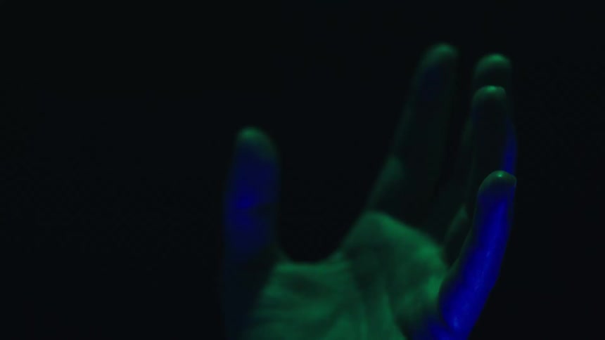 A close view shows a persons hand in a dark space with a light illuminating it in blue green and red as the hand moves and turns