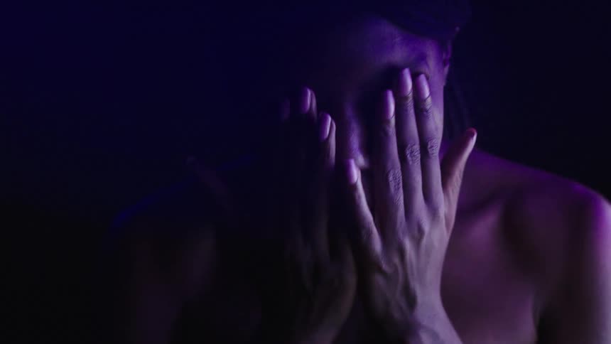 Person lit by purple light holds their face with an expression of anguish