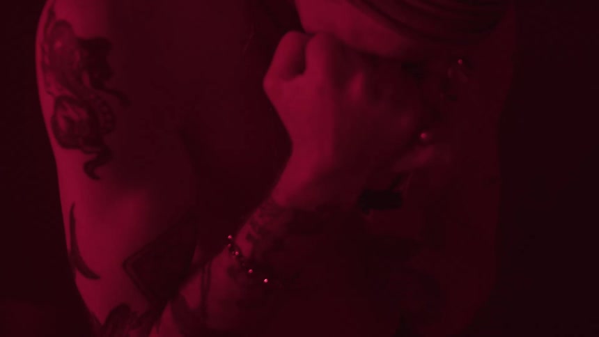 A shirtless young man with several tattoos and wearing jewelry including rings and a necklace rests his fists on his face expressing sadness in a dark room illuminated by dim red light