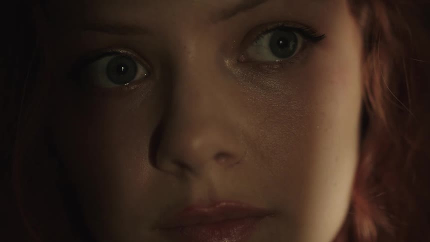 A close view shows a woman with long light brown hair and a serious expression looking directly into the camera while the left side of her face is illuminated by a beam of warm light
