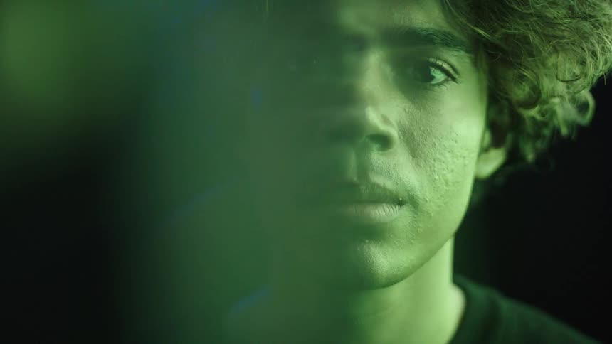 A closeup of a young mans face illuminated by shifting colorful light in a dark moody environment