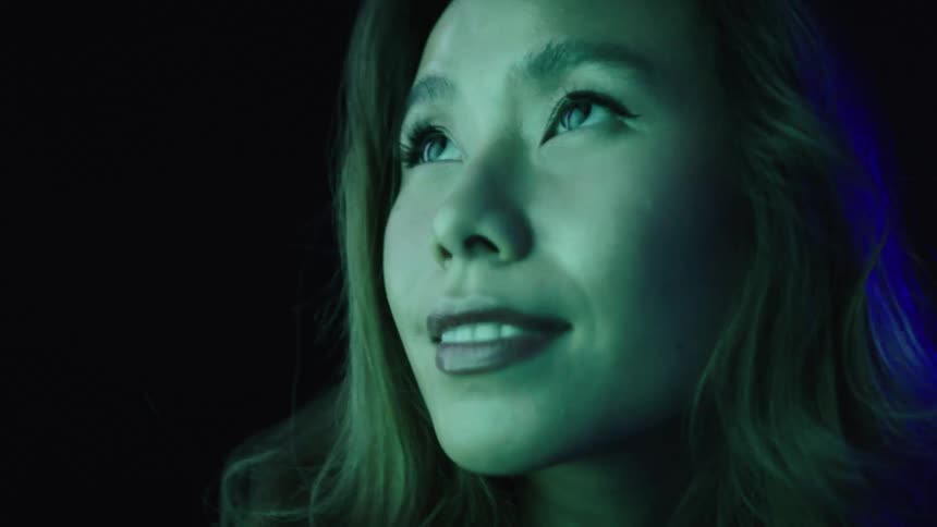 An asian woman with long hair gazes upward with a faint smile lit by green and blue light