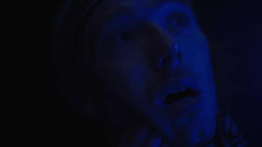 A mans face is seen in a close view with his eyes closed and his hand resting on his forehead as a blue light illuminates his face from the side