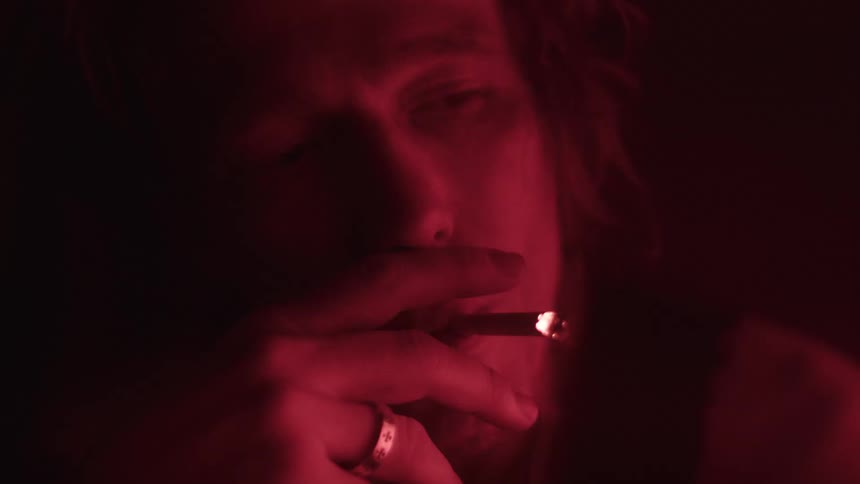 A person with long dark hair is smoking a cigarette in a dark room with their face and hands illuminated by a red light