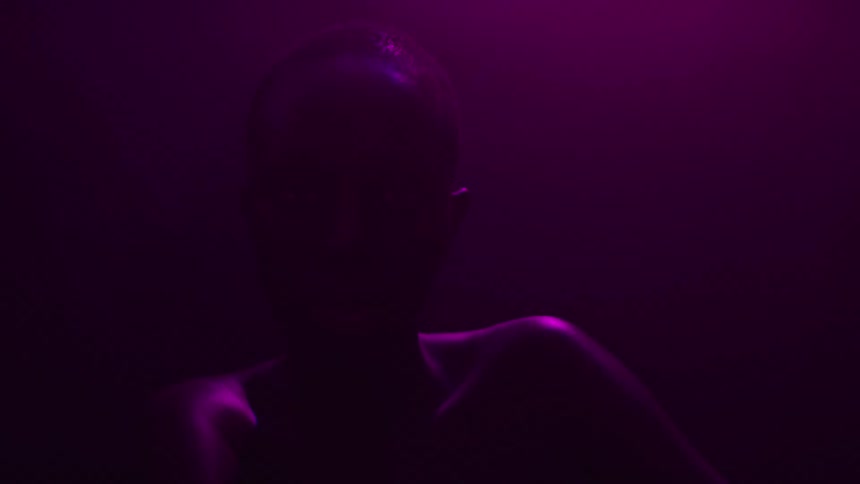 A dark silhouette of a person with a shaved head and a bare chest is visible in a dark room as they lift their hands while multicolored lights flash on them and their shoulders