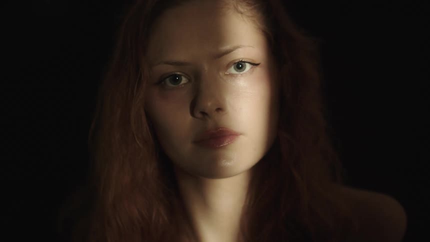 A close view shows a woman with long light brown hair and a serious expression looking directly into the camera while her face is illuminated by a beam of light