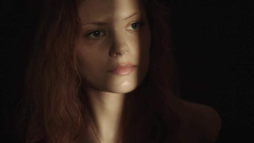 A close view shows a woman with long light brown hair and a serious expression looking off to her left as a beam of warm light illuminates the left side of her face