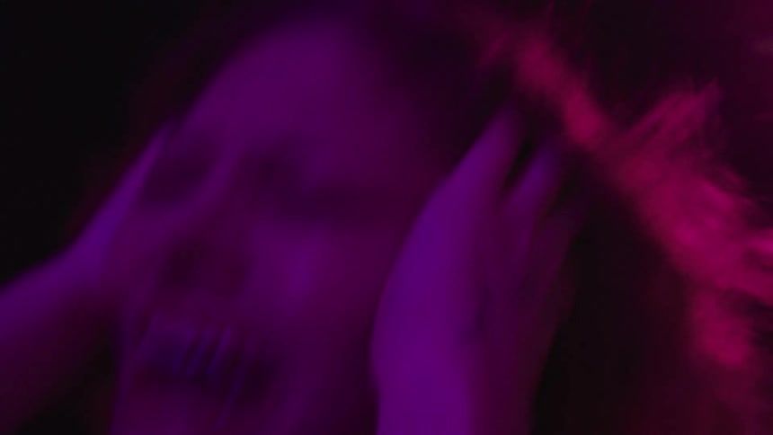 A dark and blurry view shows a woman holding her head with her hands while screaming with a purple light passing over them and a blue and red light flashing from the right