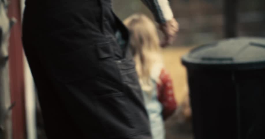 A child with long light colored hair in overalls walks with a person walking past a black garbage can