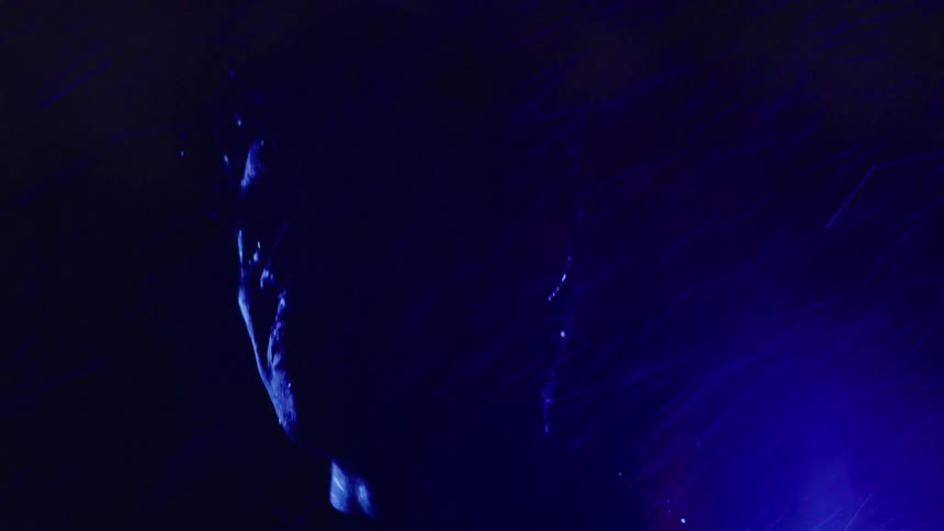 Face partially illuminated in shades of blue and purple in the dark in the rain
