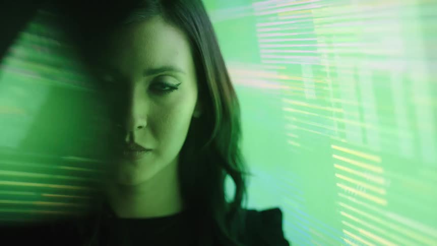 A young woman with long black hair stands beside a large screen displaying a distorted image