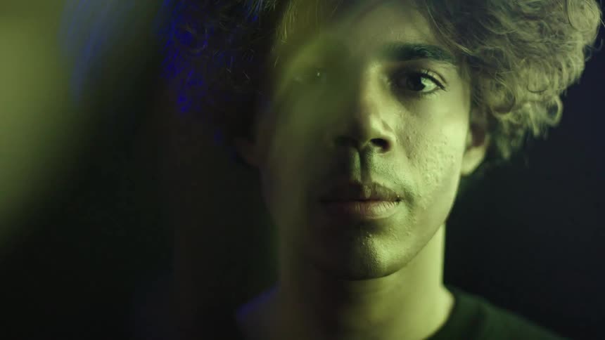 A teenager face is seen in a close view with his eyes looking directly into the camera as a colorful flashing screen illuminates the left side of his face