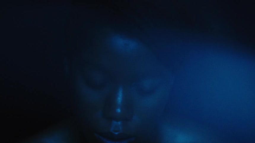 A close view shows a person with dark skin in a dark space with their face partially illuminated by a blue light as they blink and look directly into the camera