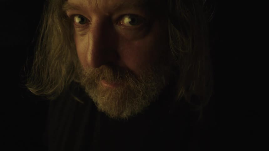 A person with a beard and long lightcolored hair is looking intently at the camera the lighting is very low casting deep shadows on their face and highlighting their eyes