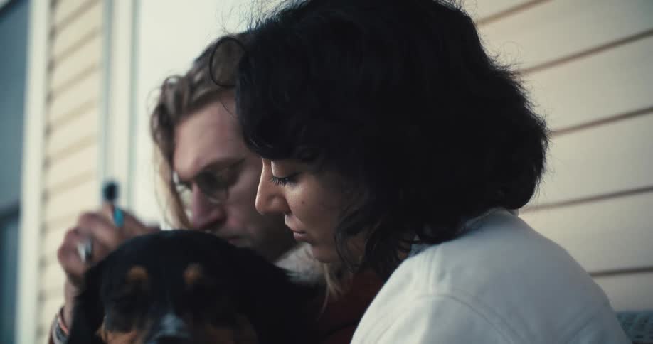 A woman with dark hair sits with a man with long light colored hair and a beard as they both pet a tri colored australian shepherd dog that is sitting between them on a porch