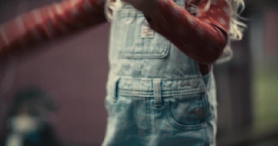 A very close view shows a child with long light colored hair and denim overalls over a red floral shirt as they hold and play with a piece of bread