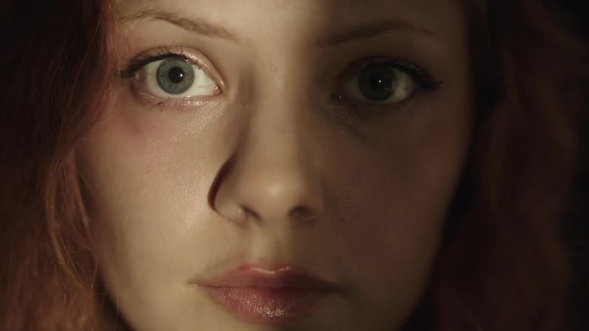 Close up of pretty white womans face in the dark as she looks into the camera and slowly blinks while a small spotlight moves back and forth horizontally across her face
