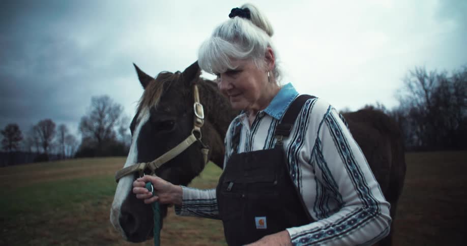 An old woman walks on a grass field pulling a horse beside her by the reins