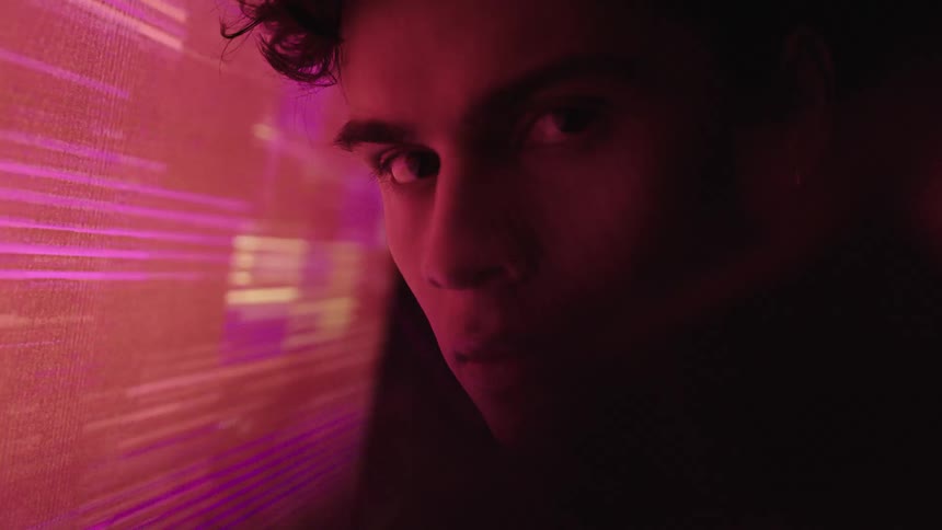 A mans face is seen in a close view with the left side illuminated by a flashing colorful screen while the right side remains in shadow as he looks directly into the camera
