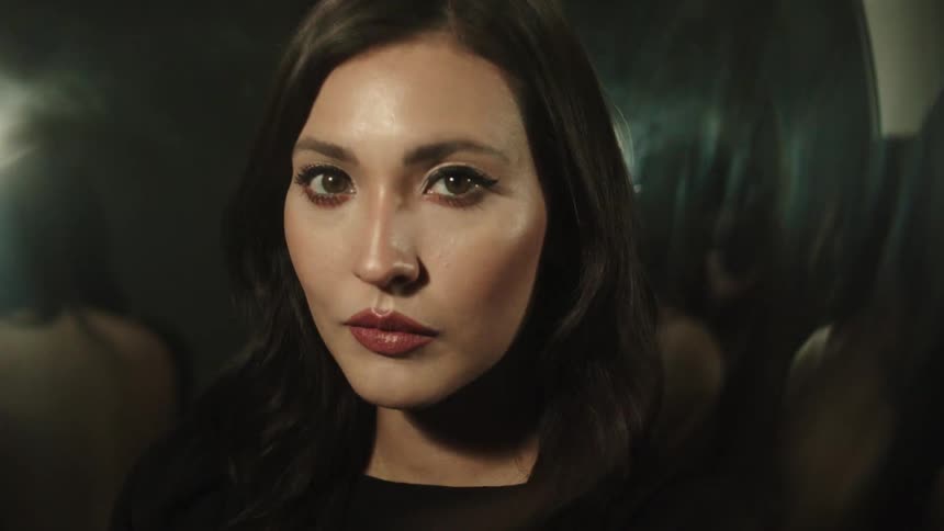 The video shows a woman with dark brown hair and makeup looking directly at the camera she blinks slowly and then turns her head slightly away
