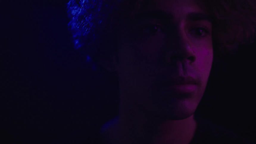 A mans face is seen in a close view with his eyes looking directly into the camera as a colorful colored light illuminates the side of his face