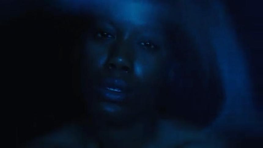 The video shows a person with a dark complexion and an expression of emotion the scene is dark with a bright blue light shining on their face