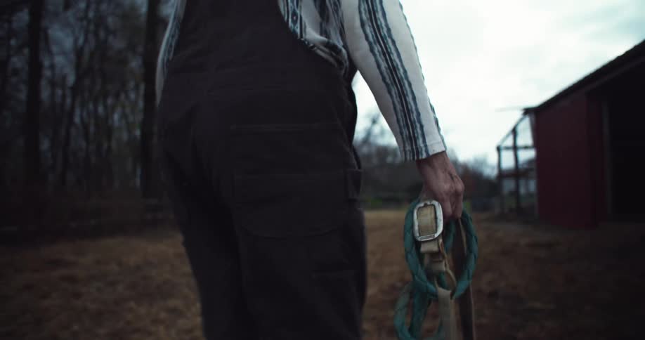 An elderly person walks towards a barn carrying horse reins