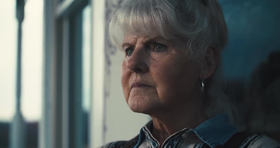 A closeup side profile of an elderly woman with grey hair and a serious expression wearing a denim shirt while she looks away on a rural setitng