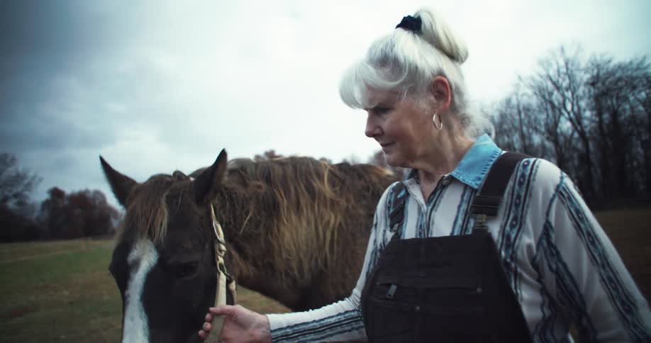 A woman with white hair is in walking in a open rural field while she is holding a horse
