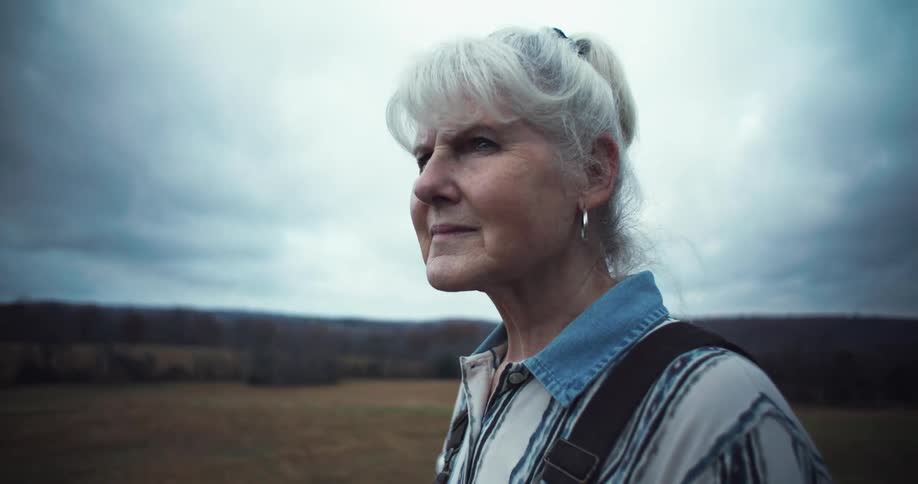 An elderly woman stands in a field and looks to the left with a pensive expression on her face