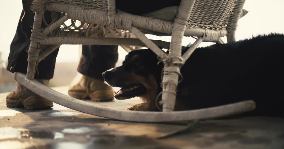 A dog with dark fur is seen lying under a persons rocking chair with its tongue hanging out as if it is panting the dogs owner is sitting in the chair wearing brown pants and boots