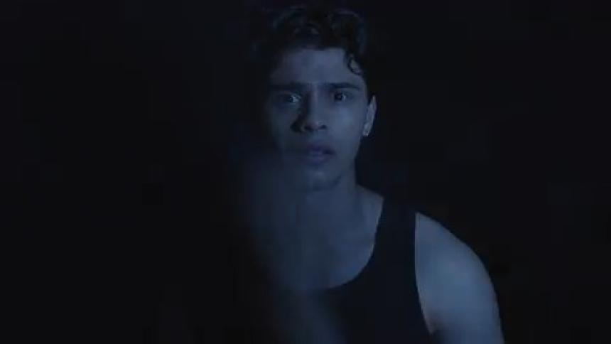 Young man in a black tank top looks forward in a dark setting lit by bluish light
