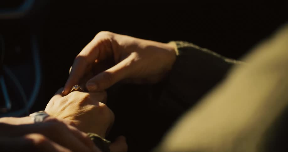 Two peoples hands with one persons hand sliding a simple silver ring onto the finger of the other against an outoffocus background inside a vehicle