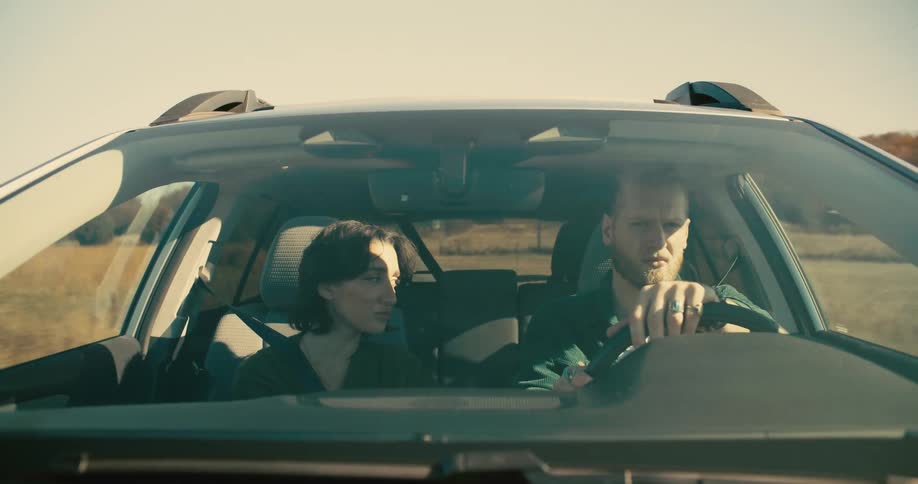 A man with light hair and a beard drives a car while a woman with dark hair sits in the passenger seat looking at him with a serious expression