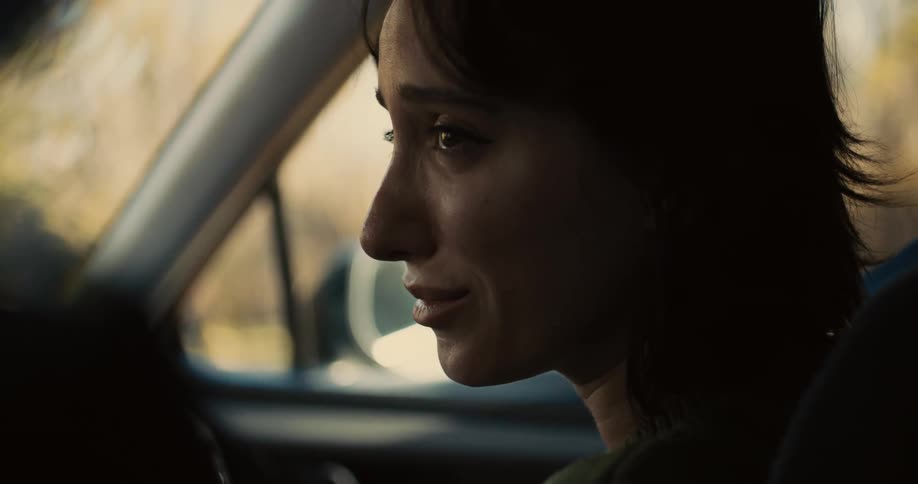 A woman with short dark hair sits in the passenger seat of a car with a sad expression as she raises her hand to her chin