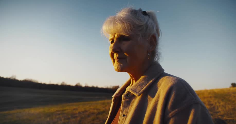 An elderly woman with her gray hair in a ponytail is standing in a field at sunset