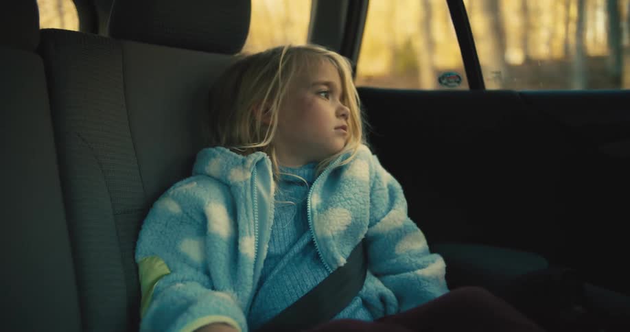 A young child with light colored hair and a blue fleece jacket sits in the back seat of a car looking out the window then rubs their eyes and yawns