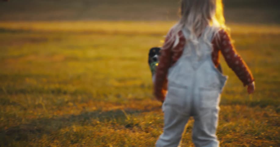 A woman and a child with long light colored hair play with a dog in a field during the golden hour