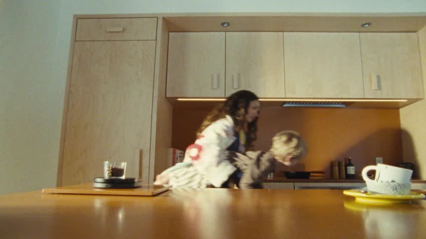 The video shows a woman with long curly hair holding a small child who is wearing a striped outfit they are in a modernlooking kitchen and the child appears to be playing with a packet of coffee beans