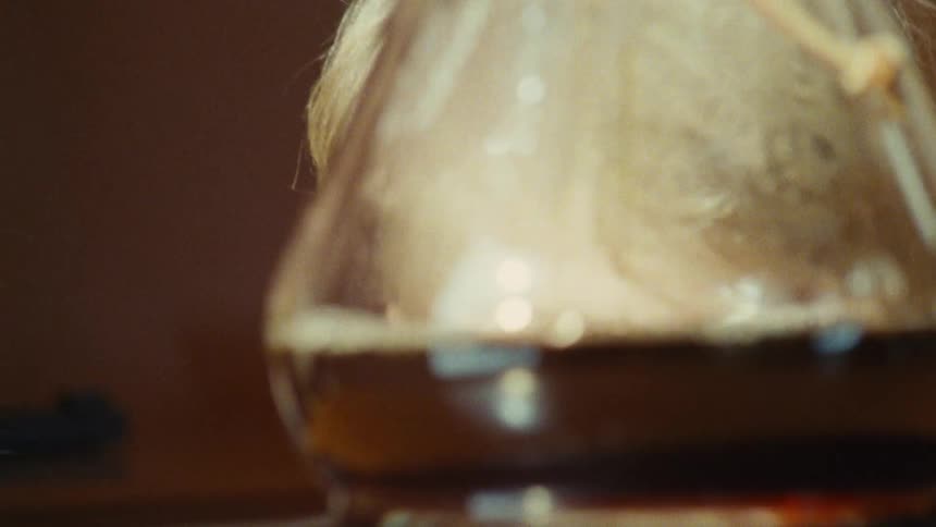 A woman and a young boy near a glass flask heating up a dark liquid with the boy looking away as the woman leans in close to the flask