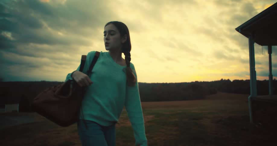 A teenage girl carrying a handbag walks across a field at sundown