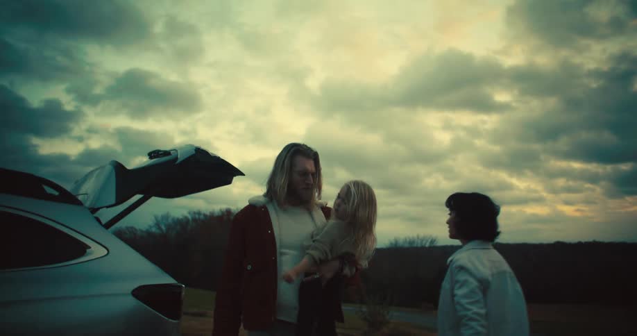 A man and a woman walking away from a car with an open car trunk holds a child with light colored hair with a cloudy sky visible behind them