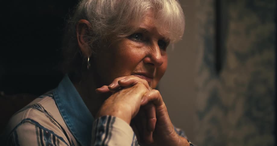 A close view shows a woman with white hair a gold hoop earring and a white and blue striped shirt sitting with her hands clasped together and a serious expression on her face