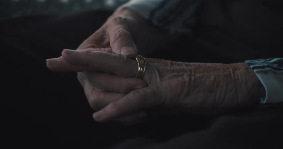 Elderly hands clasped together with a gold ring on the finger resting on the lap