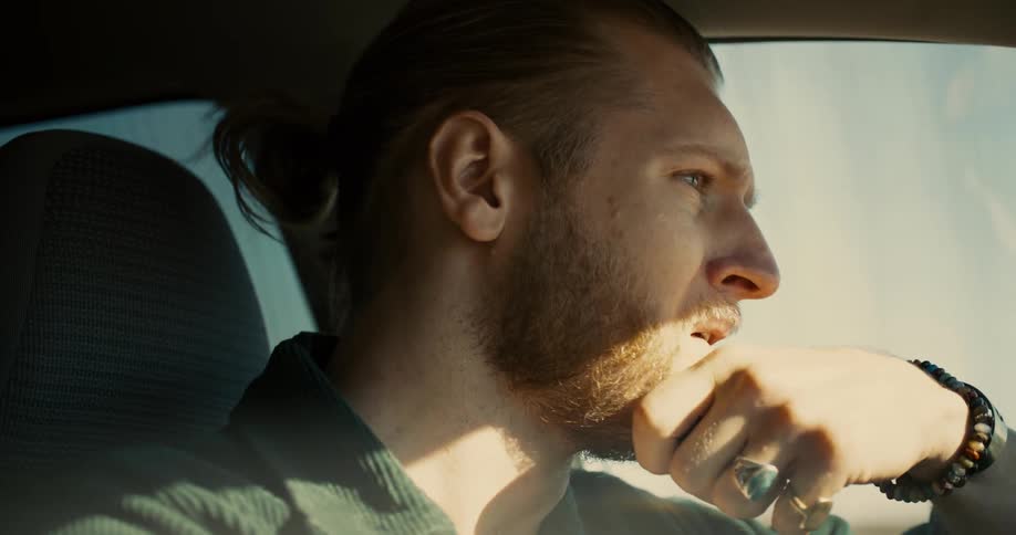 A closeup side profile shot of a man with a beard and a ponytail is shown as he is driving a car he appears to be speaking to someone offcamera and looking intently at the road ahead