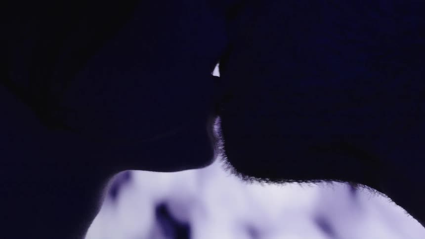 A silhouette of a couple in a dark space with a blue and white light illuminating the background as they turn their heads and lean in to kiss