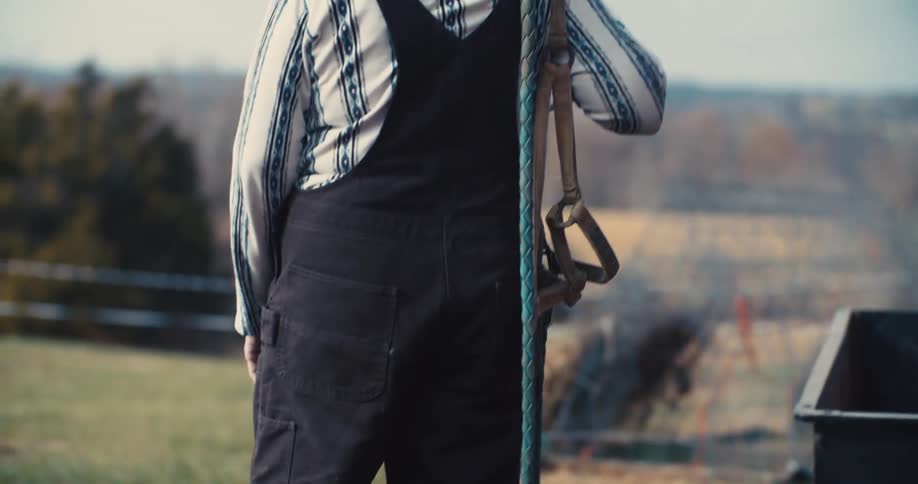 A woman is standing with her back to the camera in a rural outdoor setting wearing striped shirt and overalls and holding a stick with a rope attached